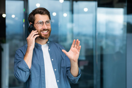 Successful smiling businessman in casual shirt talking on phone, male boss in glasses and beard near window inside modern office.の写真素材
