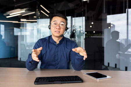 A young Asian man sits in the office at a desk in a headset, looks and talks to the camera....