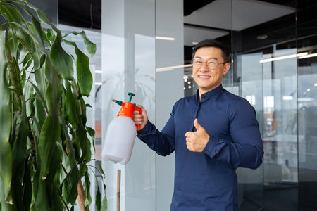 Young male office worker, programmer, freelancer. Asian takes care of plants. Waters and sprays them. He looks at the camera, smiles, points with a super finger.の写真素材