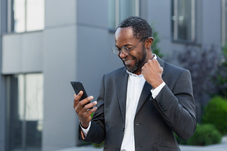 African american businessman boss outside modern office building using phone, senior man celebrating victory success reading good news online from smartphone, holding hand up gesture of triumph .の写真素材
