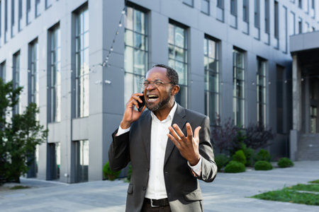 Angry african american boss arguing and shouting while talking on the phone, senior man in business suit outside office building.の写真素材