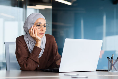 Serious bored businesswoman inside office, muslim woman in hijab thinking while sitting at workplace...
