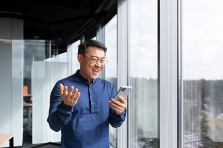 Successful asian boss near window received good news online, businessman use phone and celebrate victory and good achievement results inside office, man in shirt and glasses excitedly rejoice.の写真素材