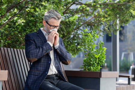 Mature businessman working outdoors with laptop, senior man sneezing from allergies, sitting on park bench on bright sunny day, boss in business suit.の写真素材