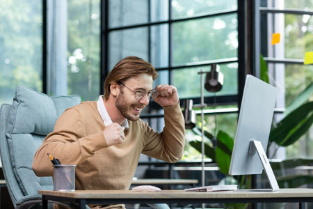 Young man in office celebrating victory and successful achievement results, businessman holding hands in triumph gesture and reading good news from computer , blond programmer in casual clothes.の写真素材