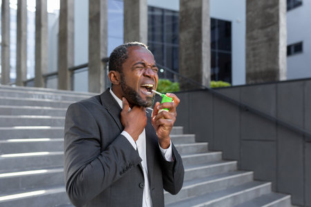 An African-American businessman is standing on the street near an office center and blowing an aerosol into his throat. He feels severe pain in the throat, cough, allergy, suffocation.の写真素材