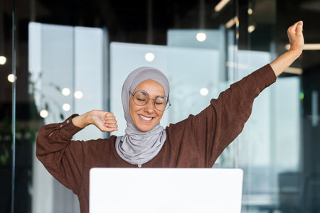 Portrait of young Arab businesswoman in hijab and glasses sitting in office at table with laptop. She is tired, has finished her work day, has done her work, stretches her hands up, smiles, rests.の写真素材