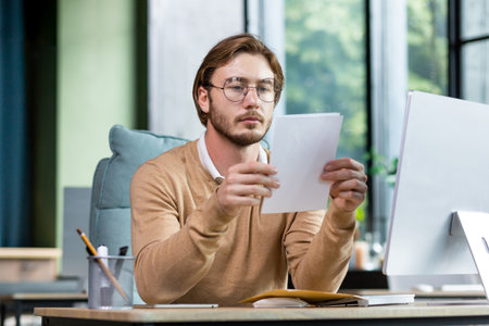 A serious young man businessman, freelancer, programmer sits at a desk in the office and reads documents, letter.の写真素材