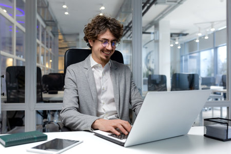 Cheerful and successful businessman inside office working with laptop, bearded man smiling and...