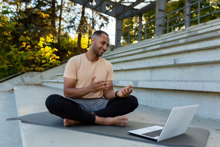 Young African American athlete fitness trainer conducts online training sitting in lotus position, man talking to physical class students using laptop and online video call.の写真素材