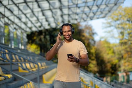 A young African-American male sportsman, athlete, runner in headphones stands in the stadium between the stands. Holds the phone in his hands, listens to music. He closed his eyes, resting.の写真素材