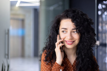 Hispanic business woman with curly hair close up talking on phone, woman inside office at workplace...