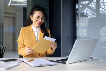 Successful hispanic woman inside office at work businesswoman received postal envelope letter notification with good news, female worker celebrating victory and good results achievement at workplace.の写真素材