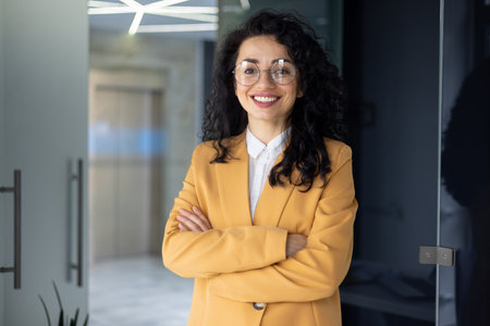 Portrait of successful businesswoman inside office, Hispanic with curly hair and glasses smiling and looking at camera, boss woman in yellow jacket, financier happy with achievement results at work.の写真素材