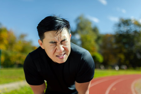 Close-up of sportsman having severe headache and dizziness, asian man in stadium doing fitness and active exercise, male runner in sportswear overworked by training on sunny day.の写真素材
