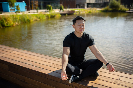 Asian young male athlete, trainer, practitioner sits by the lake on a bench in the lotus position. He closes his eyes, meditates, does yoga, rests.の写真素材