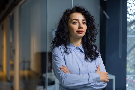 Portrait of serious female boss inside business company office, businesswoman crossed arms looking concentrated at camera, wearing shirt, satisfied and successful hispanic woman with curly hair.の写真素材
