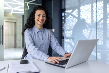 Portrait of successful hispanic woman at workplace inside office, woman with video call headset smiling and looking at camera, support worker working with laptop, help desk.の写真素材