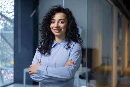 Portrait of successful business woman inside office at workplace, hispanic woman satisfied with achievement smiling and looking at camera at work with arms crossed boss in shirt on company corridor.の写真素材