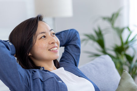Closeup photo of a young beautiful Asian at home in the living room, woman has her hands behind her head and is resting, breathing fresh air, smiling and looking window, dreaming happy future.の写真素材