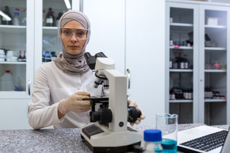 Portrait of young Muslim woman in hijab, scientist, chemist, biologist sitting at a table with a microscope in a research center. He looks seriously and intently at the camera.の写真素材