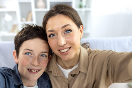 Son and mother at home sitting on the sofa in the living room, looking at the camera, smiling, taking a selfie photo, and talking via video call online.の写真素材