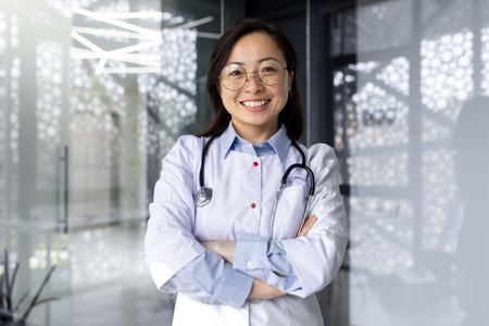 Portrait of a young beautiful and successful Asian woman inside a clinic office, a doctor smiling and looking at the camera with crossed arms and a white medical coat with a stethoscope.の写真素材