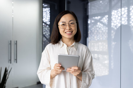 Portrait of successful asian female programmer inside office, young woman smiling and looking at camera, using tablet computer to test application software, workplace inside office.の写真素材