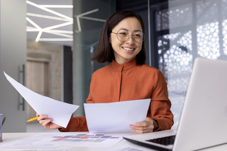 Young successful asian financier behind paper work inside office, businesswoman accountant reviewing contract documents and reports smiling and happy, satisfied with achievement results using laptop.の写真素材