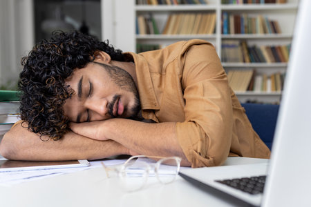 Tired student sleeping on table inside university academic library, hispanic man among books with laptop preparing for session and final exams.の写真素材