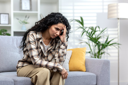 Lonely hispanic woman sitting alone on sofa in living room at home, depressed woman thinking about difficult decision.の写真素材