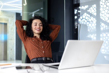 Happy thinking woman at workplace inside office, business woman with hands behind head completed work well, latin american woman satisfied with achievement results looking at laptop screen.の写真素材