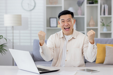 Portrait of successful Asian businessman mature man working remotely from home freelancer, living room sitting at table looking at camera and happy holding hands up celebrating victory and triump.の写真素材