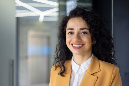 Closeup portrait of mature adult business woman, boss smiling and looking at camera, pleased hispanic woman with curly hair inside office at workplace.の写真素材