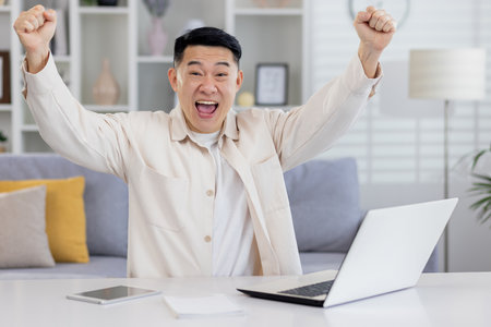 Portrait of successful Asian businessman mature man working remotely from home freelancer, living room sitting at table looking at camera and happy holding hands up celebrating victory and triump.の写真素材