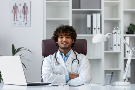 Portrait of a successful Hindu doctor inside the office of a modern clinic, sitting at the workplace at the table smiling and looking at the camera with his arms crossed, a man in a white coat.の写真素材