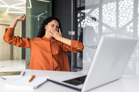 Tired businesswoman in office stretching and yawning, latin american woman indoors working at workplace using laptop bored overtired.の写真素材