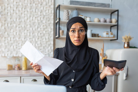 Young worried Muslim woman in hijab holding documents and calculator. Financial problems with accounts, loans, family budget. He looks sadly at the camera.の写真素材
