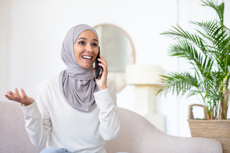 Young arab woman in hijab sitting on sofa at home and talking on the phone. Gesturing with hands....