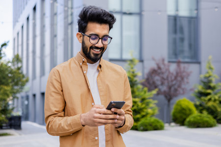 Successful Indian young businessman outside office building walking in daytime, man holding phone in hands, businessman dialing, browsing online pages, programmer engineer in glasses.の写真素材