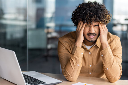 Tired young businessman working inside office at workplace, hispanic man has severe headache stress and overload, man sitting at workplace and leaning hands on head.の写真素材