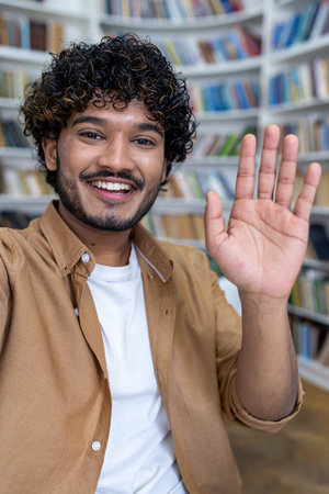 A young happy Spanish male student is talking on a video call from a phone in the library. Greets...