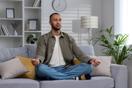 Yoga classes at home. Young African American man sitting relaxed on couch in lotus position with eyes closed, meditating, resting.の写真素材