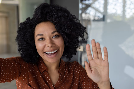 Beautiful business woman with curly hair wearing looking at smartphone camera, waving her hand in...