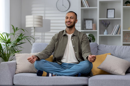 Resting at home after work. Young smiling African American man sitting on sofa in lotus position with closed eyes, relaxing, meditating.の写真素材