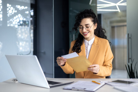Businesswoman received happy news letter from bank, Hispanic woman with curly hair and glasses works inside modern office building, uses laptop.の写真素材
