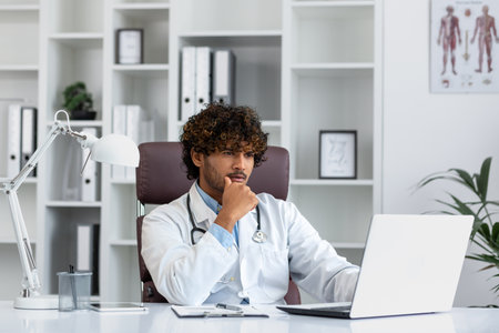Serious focused hispanic doctor working inside medical office, man in medical coat thinking looking at laptop.の写真素材