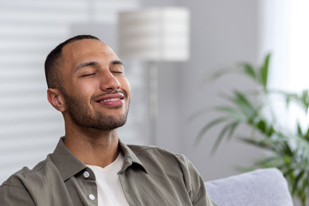 Portrait of the face of a smiling young Latin American man thoughtfully sitting at home on the sofa with closed eyes, resting, dreaming.の写真素材