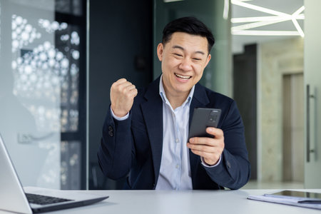 Successful asian businessman received good news notification online, workplace boss holding hand up celebrating successful achievement and winning.の写真素材