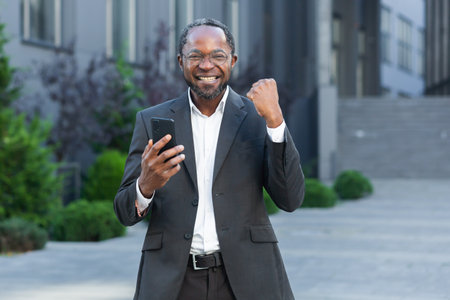Portrait successful and satisfied african american businessman boss holds phone in hands smiling and looking at camera, man received notification of successful win, holds hand up celebrating triumphの写真素材